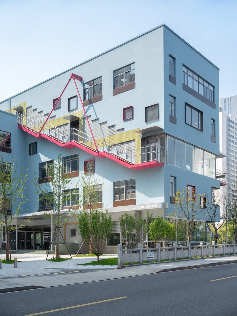 Corner facade showing the pale blue cladding, yellow tile band, and pink steel staircase with young street trees