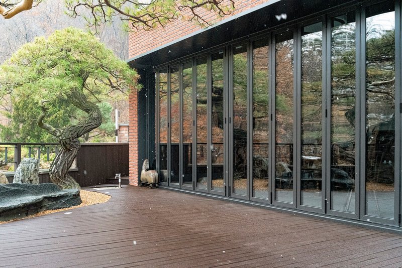 Timber deck with floor-to-ceiling glass doors beside a gnarled pine tree and stone sculpture