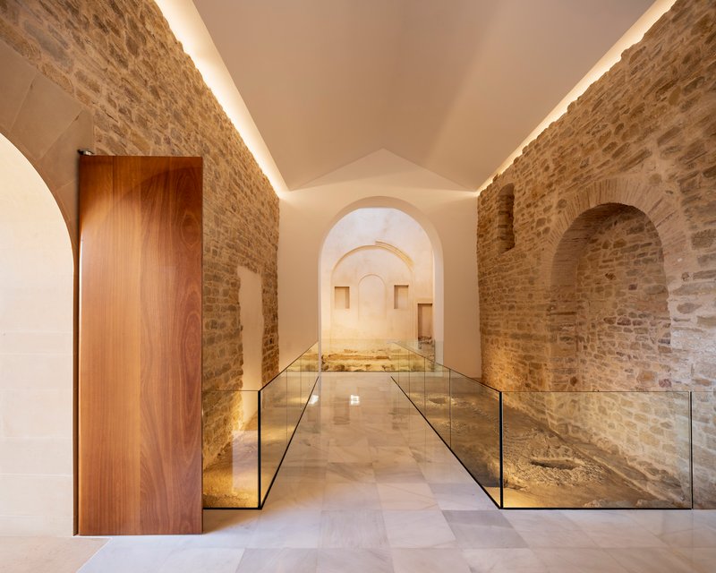 Vaulted corridor with exposed stone walls, walnut door and glass-edged floor revealing archaeological remnants below