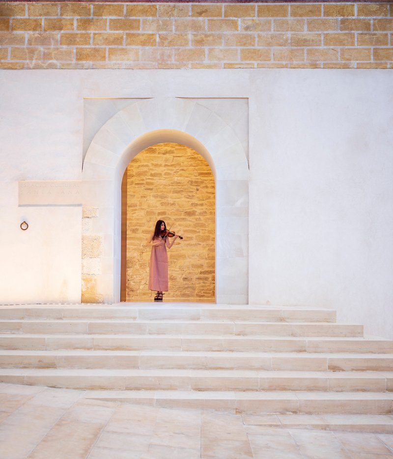 Wide stone steps leading to an arched opening in a white plaster wall with a violinist standing inside