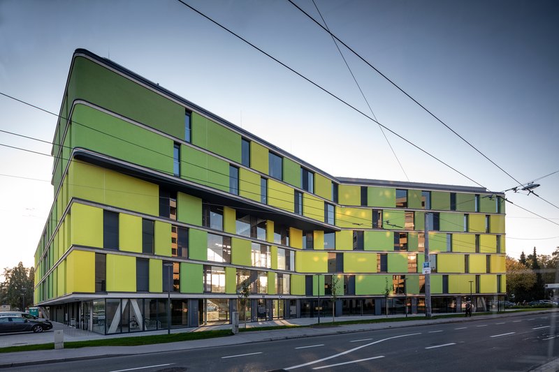 Long facade showing the chequered yellow and green panel composition with ground-level pilotis beneath a cloudy sky