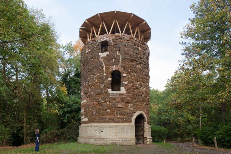 Cylindrical brick tower with zigzag timber roof structure framed by trees on a grassy clearing