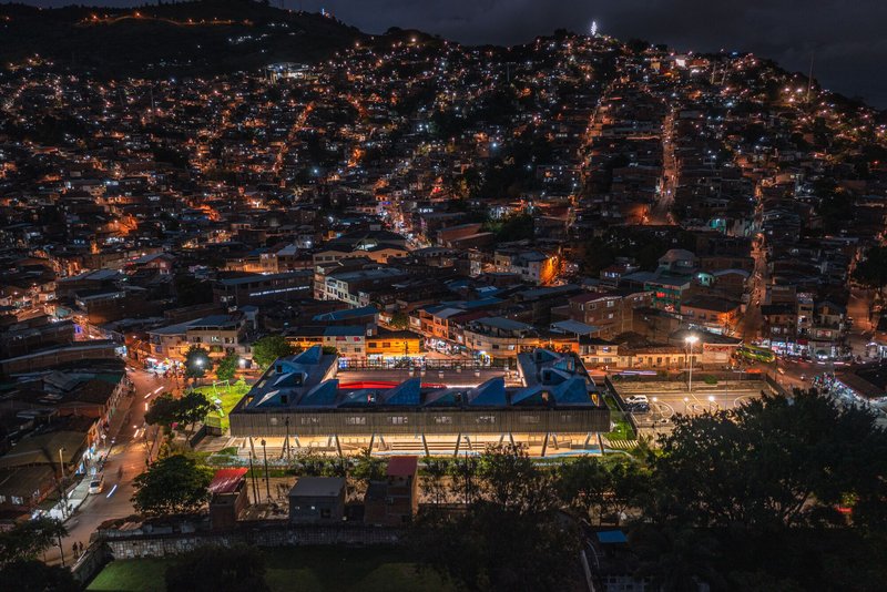Nighttime aerial view of illuminated compound with blue roof volumes nestled in hillside residential district