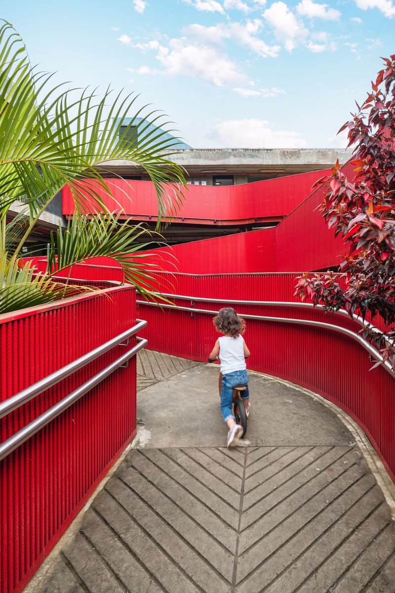 Child walking on the curved red metal ramp framed by tropical foliage under blue sky