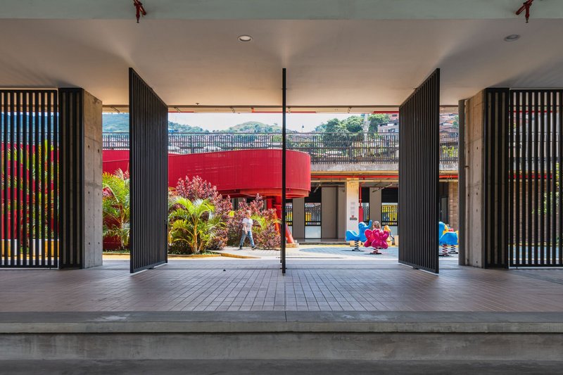 Covered pavilion with vertical slat screens framing a view of the courtyard with red walls and children playing
