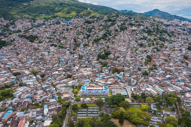 Wide aerial view of the complex with blue roofs set within the hillside neighborhood and surrounding mountains