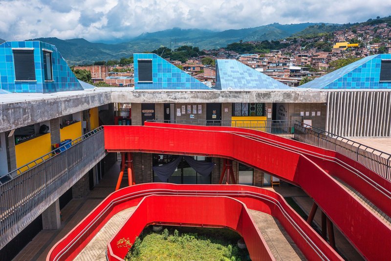 Aerial view of red curved pedestrian bridges connecting concrete buildings with blue pyramid roofs against a mountainous backdrop