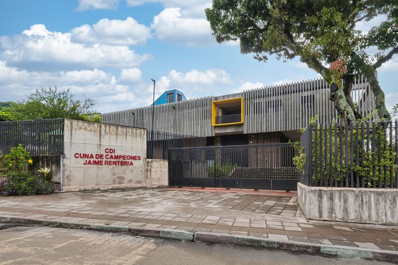 Street view of vertical metal slat facade with yellow window frame and blue pyramid roof beside weathered concrete wall