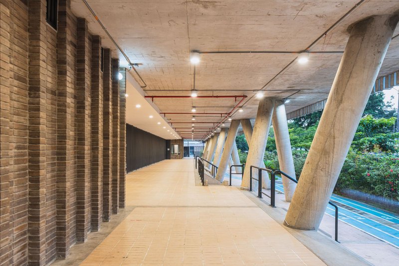 Covered walkway with exposed concrete ceiling, rhythmic brick piers, and angled concrete columns overlooking greenery