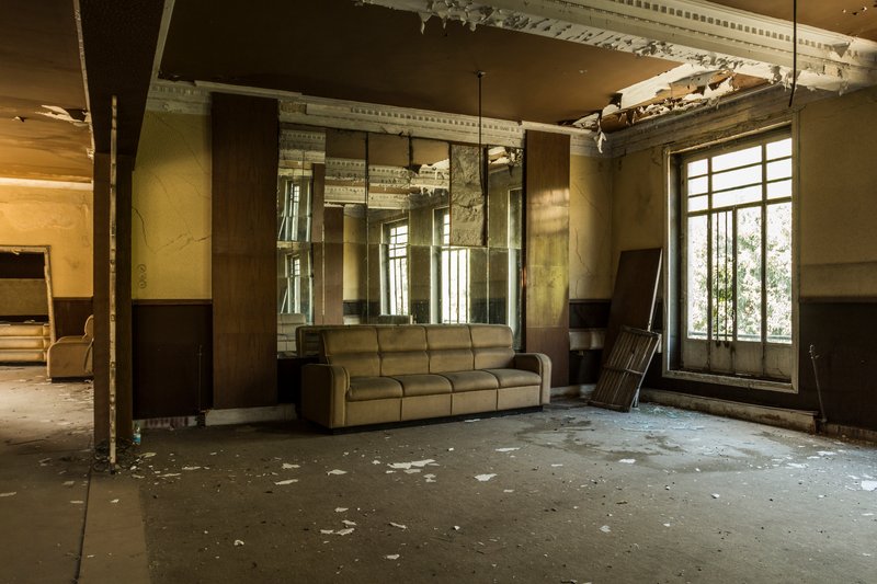 Abandoned interior hall with ornate ceiling moldings, tall windows, and a worn sofa under natural light