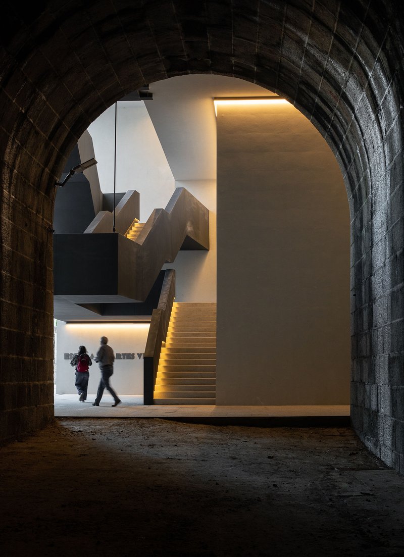 View through barrel-vaulted granite passage toward illuminated staircase with two visitors walking
