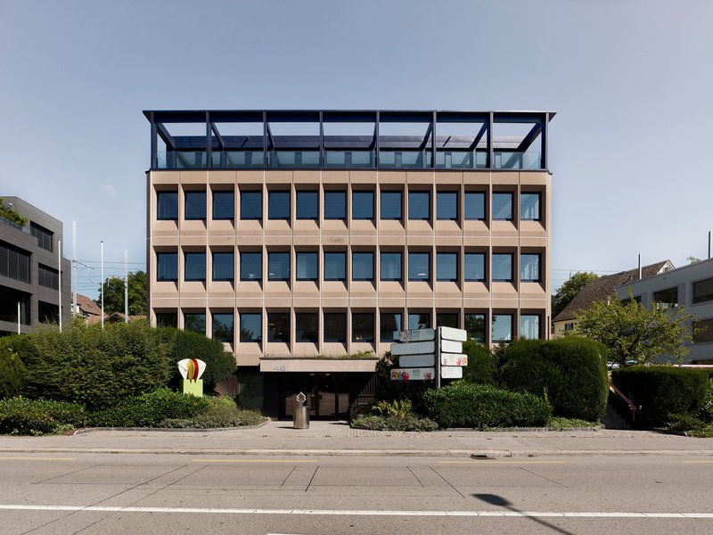 Street view of the layered facade with terracotta panels and a recessed glazed penthouse above green hedges