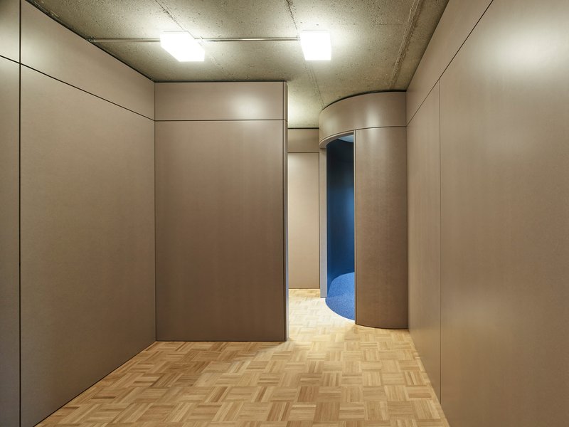 Corridor with beige wall panels, parquet flooring, and curved partition enclosing a blue space