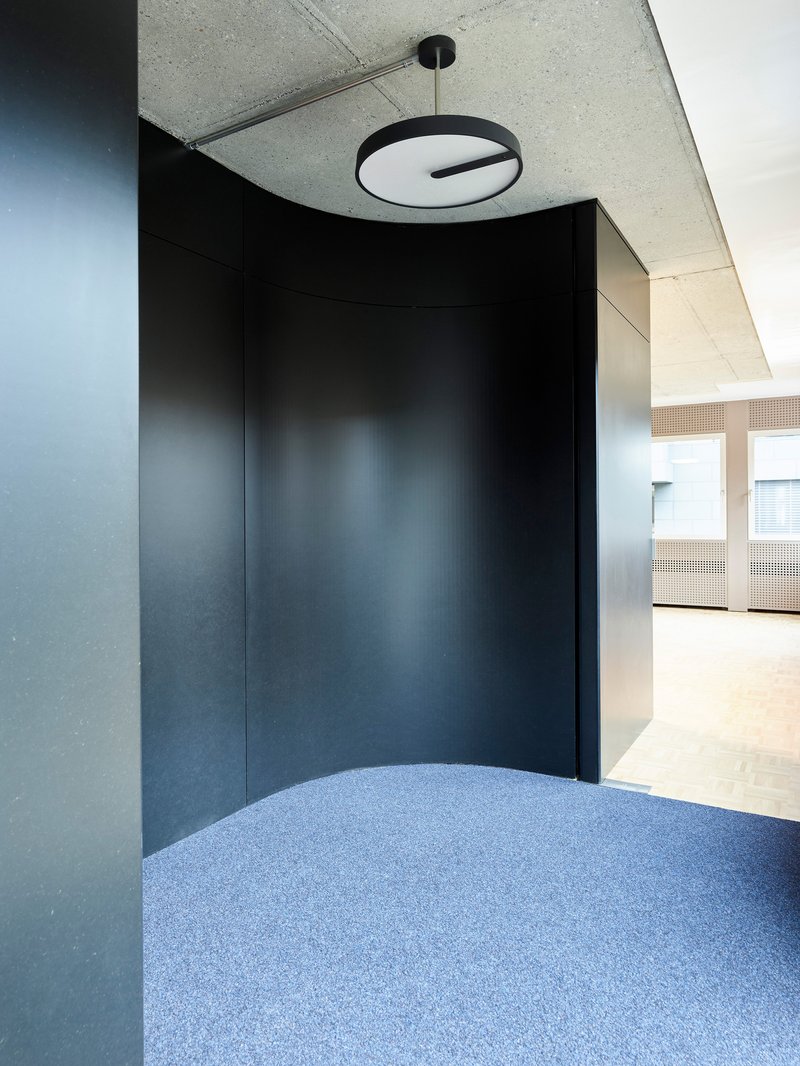 Curved dark wall meeting blue speckled flooring beneath a circular pendant light and acoustic tile ceiling