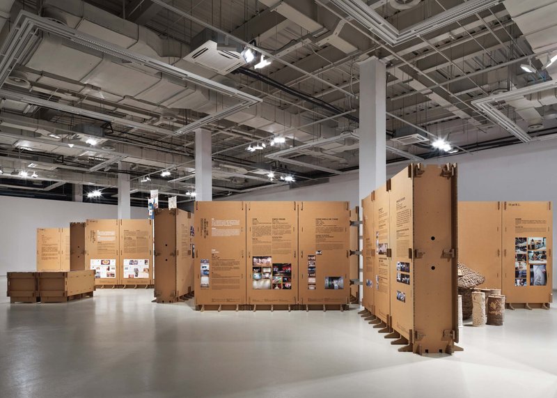 Cardboard exhibition walls arranged in a serpentine configuration with cylindrical woven elements visible at right