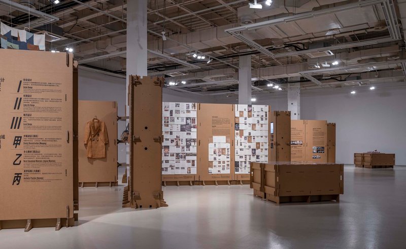 Exhibition space with cardboard panel partitions displaying photographs and text under an exposed ceiling grid