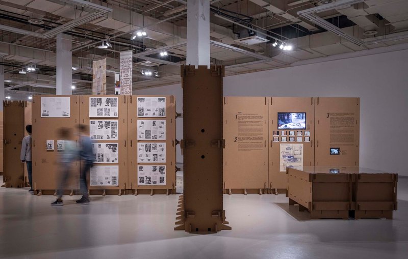 Cardboard column wrapping with stacked base elements and visitors moving through the exhibition space