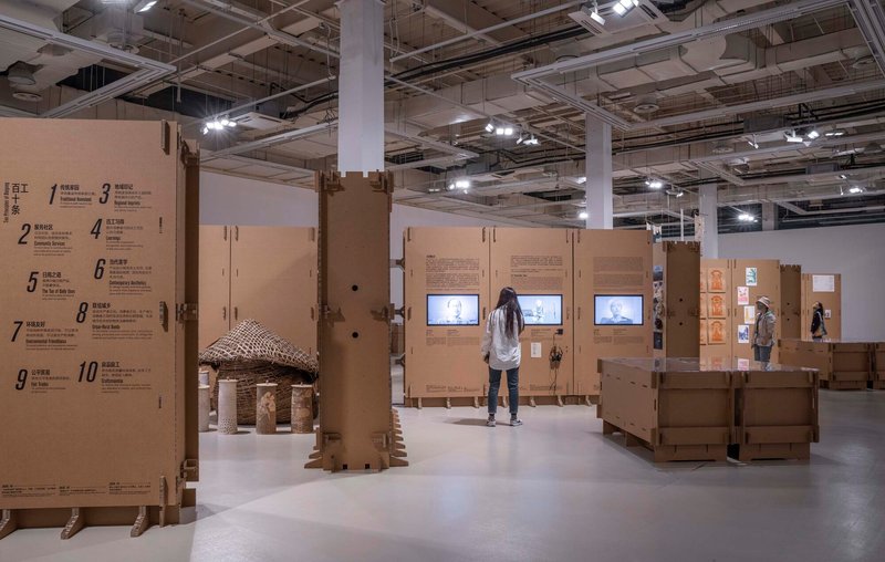 Gallery view showing cardboard display walls with embedded video screens and a small pitched-roof structure