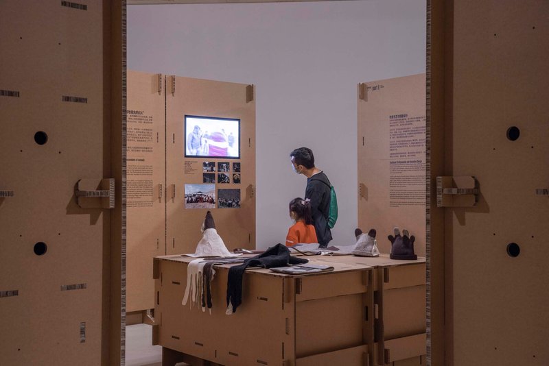 Two visitors seated on a cardboard bench viewing a video monitor between flanking text panels