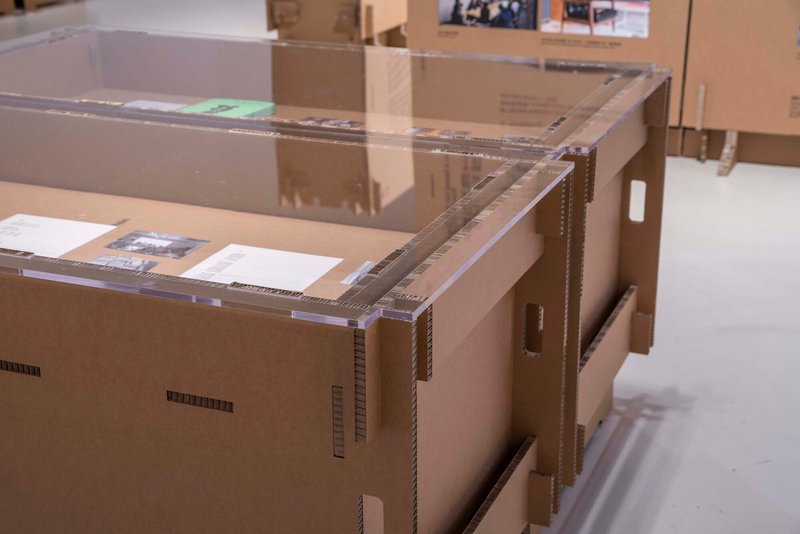 Acrylic-topped cardboard model on a white floor beneath overhead gallery lighting