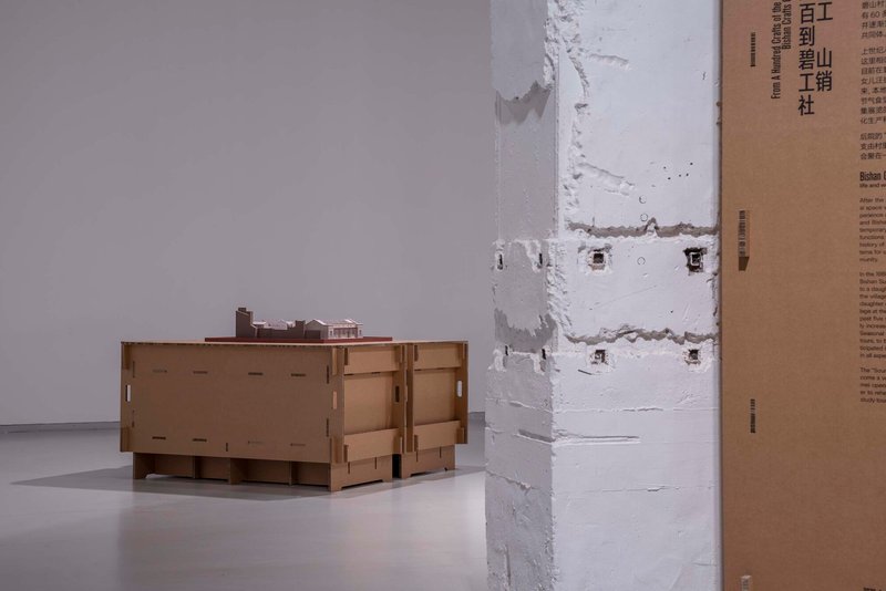Cardboard model beside a white-painted column with exposed anchor holes in a gallery setting