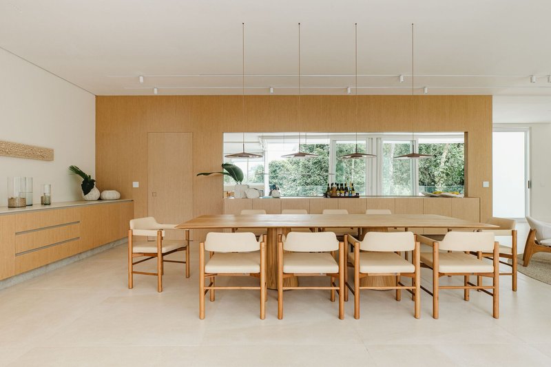 Dining room with long timber table and wood-paneled wall framing a horizontal window to greenery