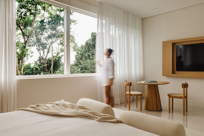 Bedroom with a figure in motion near the corner window overlooking dense green foliage