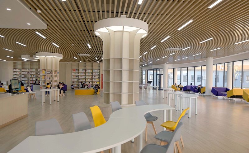 Library interior with slatted timber ceiling, white columns and scattered seating with yellow and grey chairs