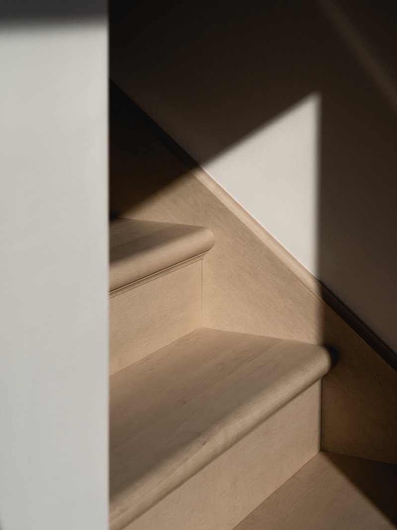 Light wood staircase with black metal handrail and white walls casting triangular shadows