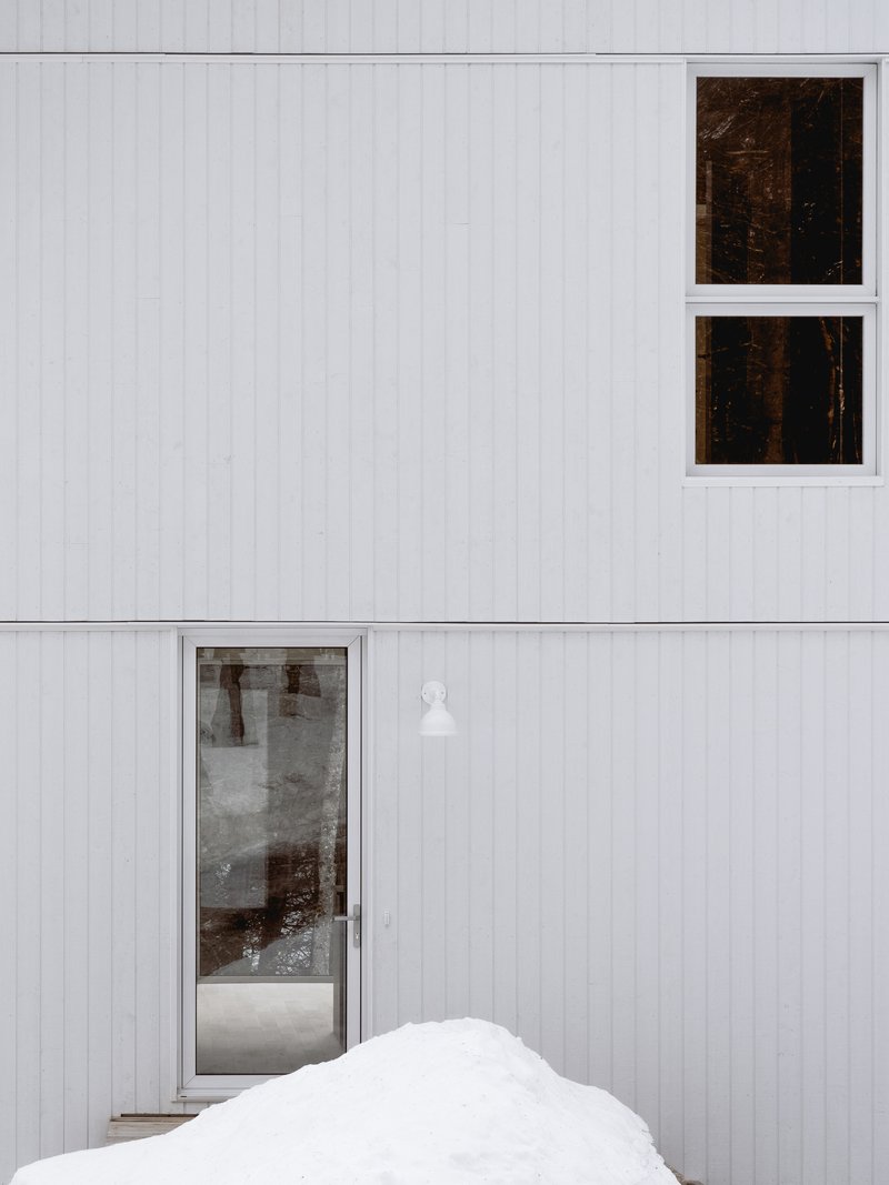 Vertical white siding with square windows reflecting forest and glazed entry door beside a snowdrift