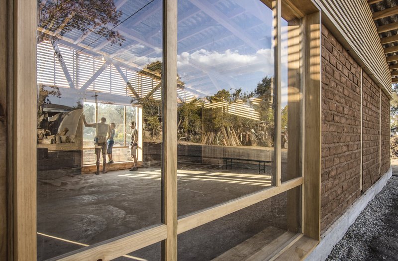 Glass-walled corridor with timber frame and slatted roof overhang casting striped shadows on concrete floor