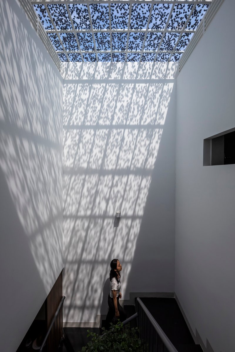 Double-height white stairwell with perforated skylight casting dappled shadows on the wall where a woman stands