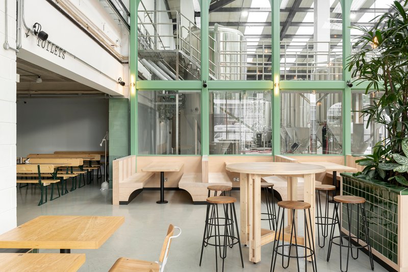 Interior dining space with pale timber booth seating and green-framed glass partitions revealing industrial brewery equipment beyond