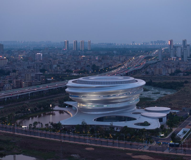 Aerial view of the spiraling ribbed volume beside a highway at dusk with city lights illuminating the background