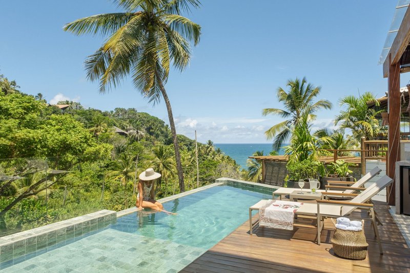 Infinity pool deck with timber loungers overlooking palm trees and the ocean beyond under clear skies