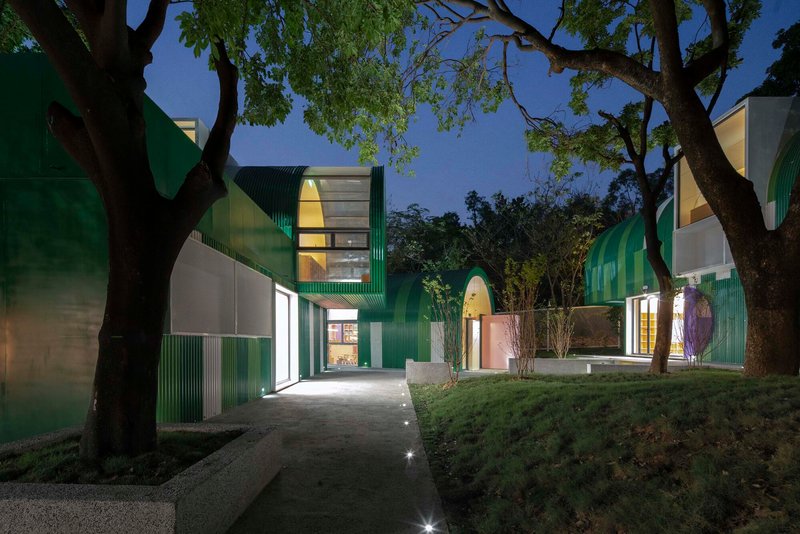 Illuminated pathway between green paneled volumes under mature trees at twilight