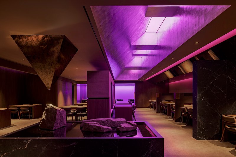 Restaurant dining hall with purple-lit ceiling panels and a rough stone sculpture centerpiece on a black plinth