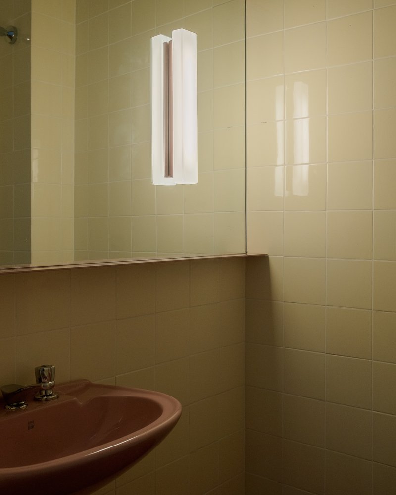 Yellow tiled bathroom corner with mirror and wall-mounted tubular light fixture above a pink sink