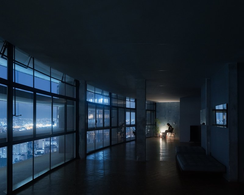 Interior space at night with figure seated beside floor lamp near glass facade overlooking city lights