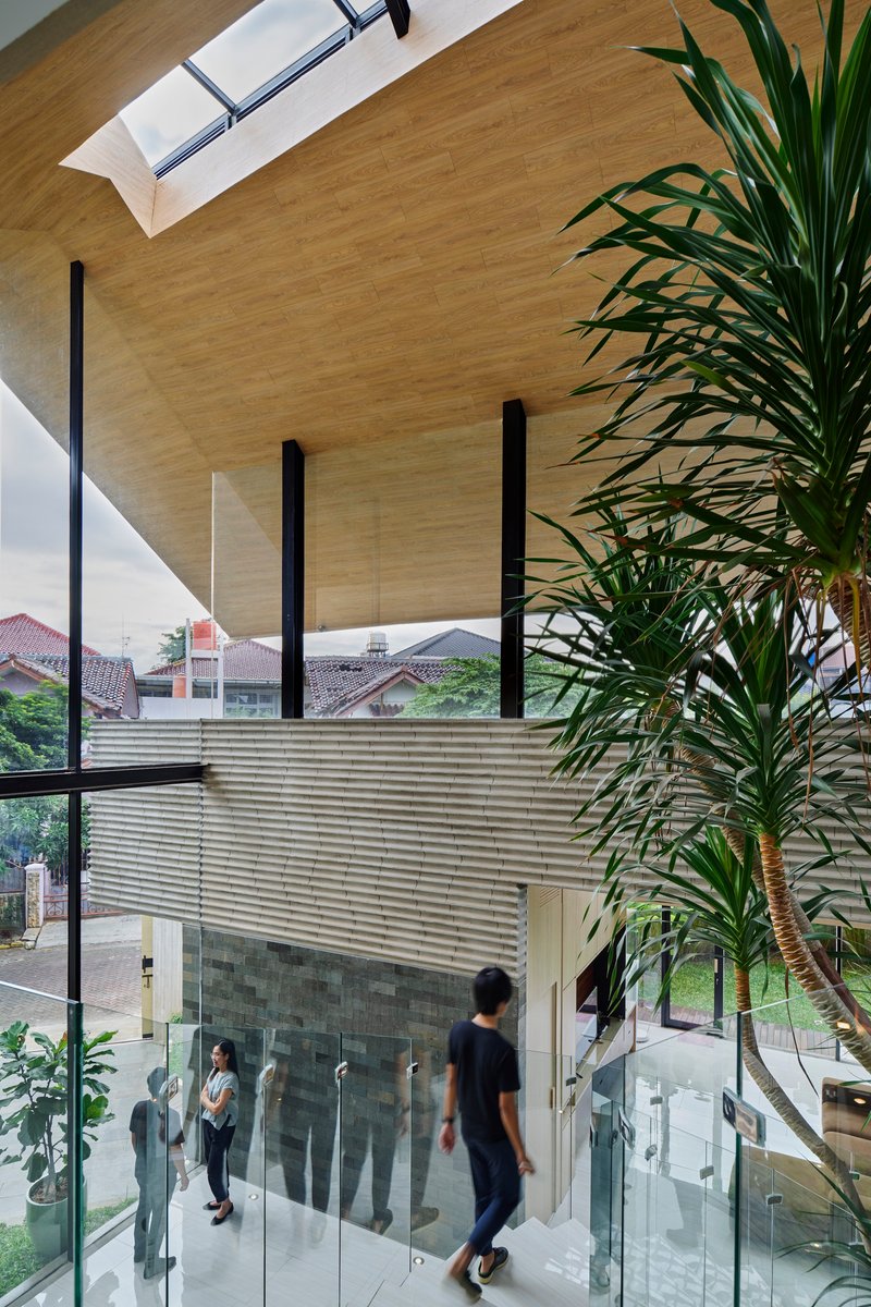 Double-height interior with plywood ceiling, black steel columns, glass balustrade and potted palms beside person descending