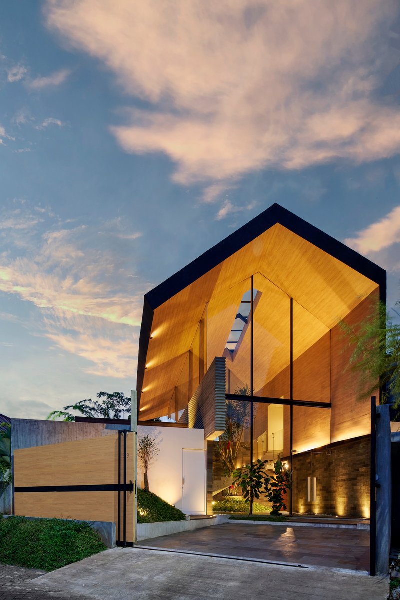 Street view of the timber-clad gabled volume illuminated at dusk with a sliding gate entry