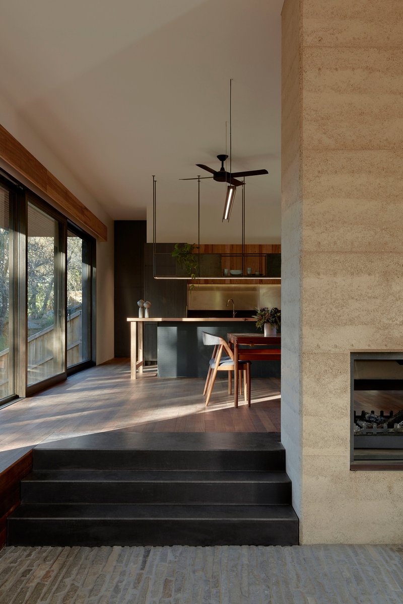 Black steel steps leading to open kitchen with rammed earth wall and morning sunlight