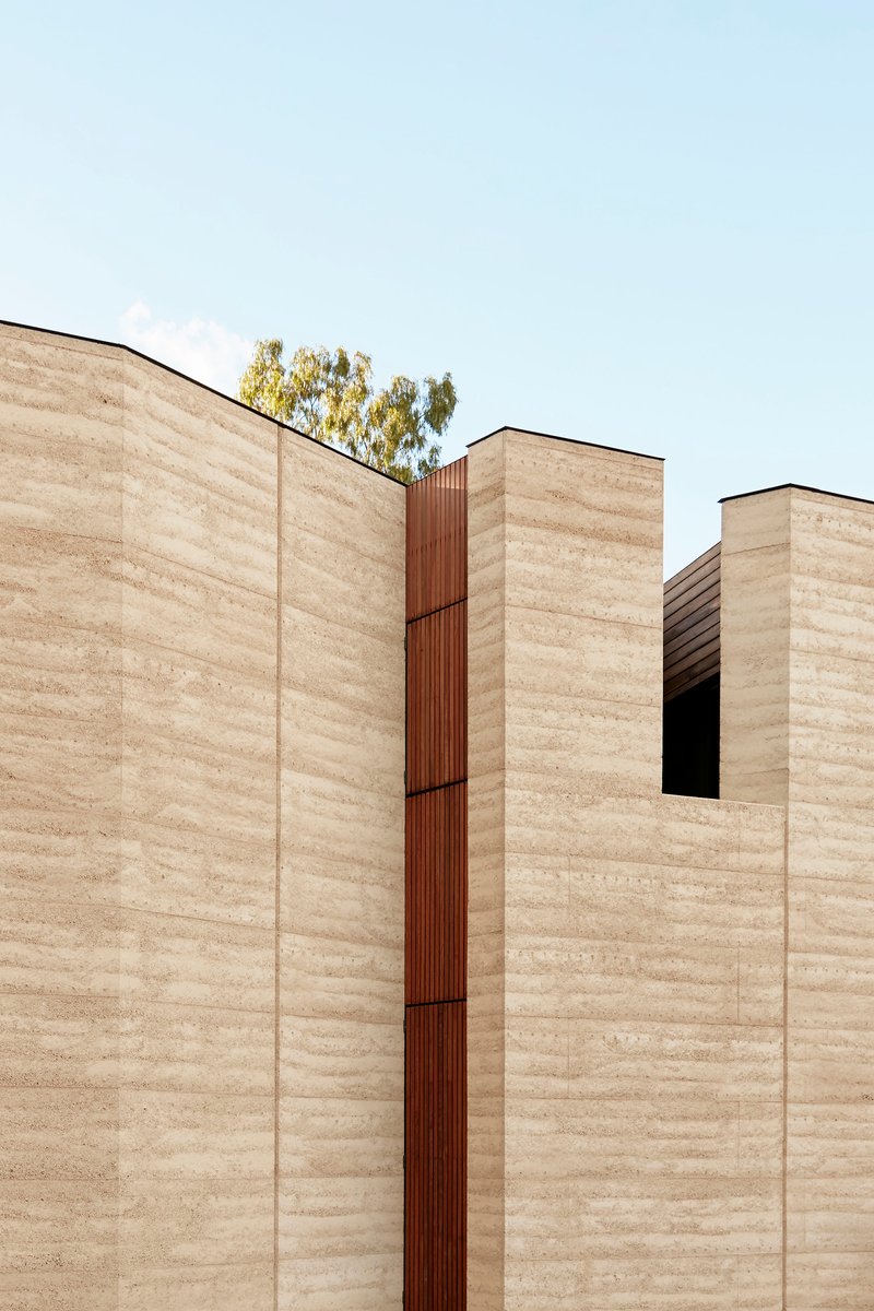 Rammed earth facade with vertical timber slot detail beneath a pale sky