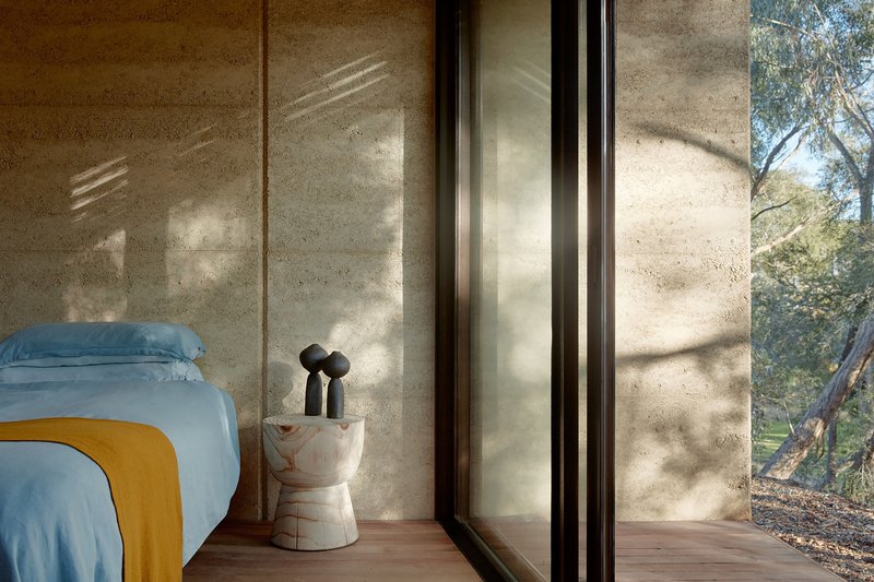 Bedroom with floor-to-ceiling glazing and rammed earth walls casting dappled afternoon shadows