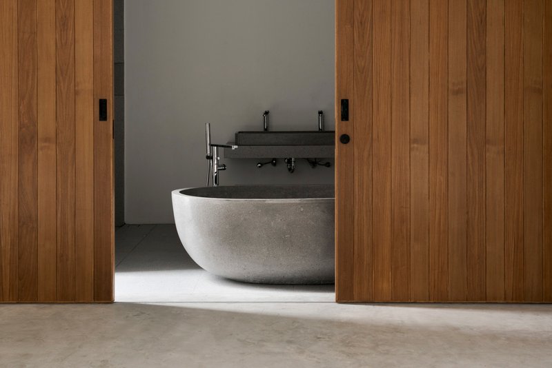 Freestanding stone bathtub framed by timber sliding doors with soft daylight across polished concrete floor