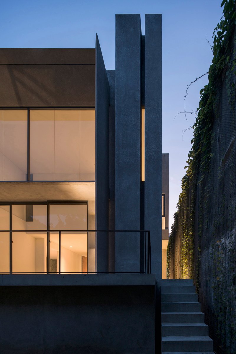 Rear courtyard showing stacked glass volumes beside a tall chimney and illuminated vertical wall planting at dusk