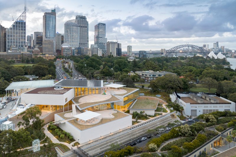 Aerial view of the terraced white volumes with golden glass walls nestled among trees and parkland