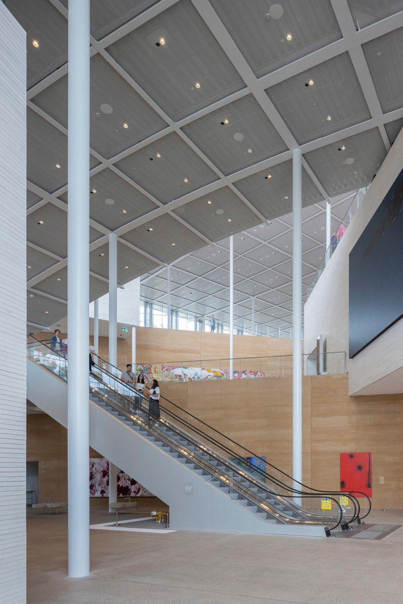 Interior atrium with escalators rising between white columns under a grey coffered ceiling with recessed lighting