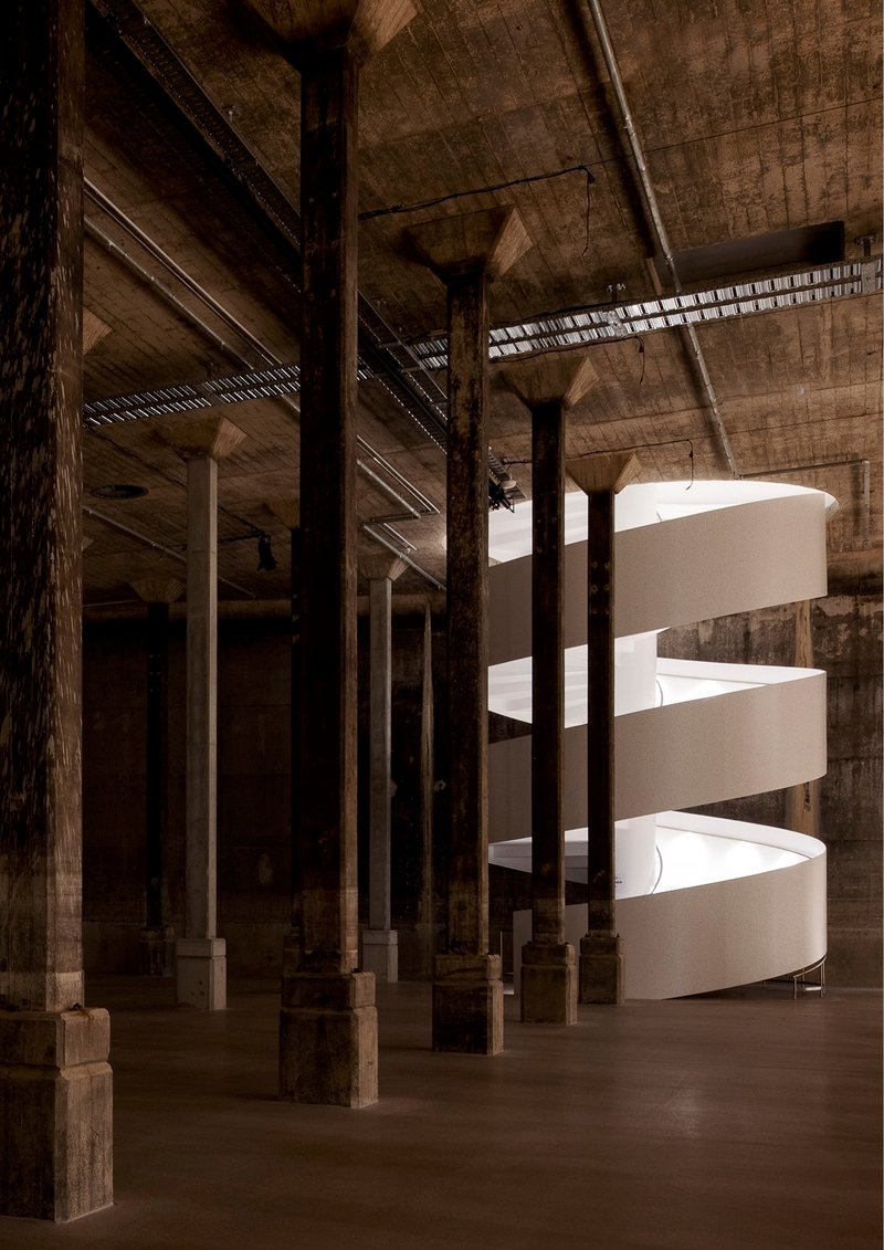 White helical staircase illuminated by clerestory slot within a raw concrete hall with timber columns