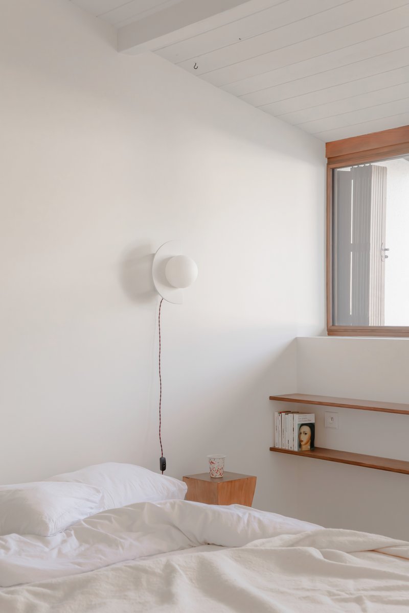Bedroom with timber window frame, white plank ceiling and wall-mounted disc sconce above bed
